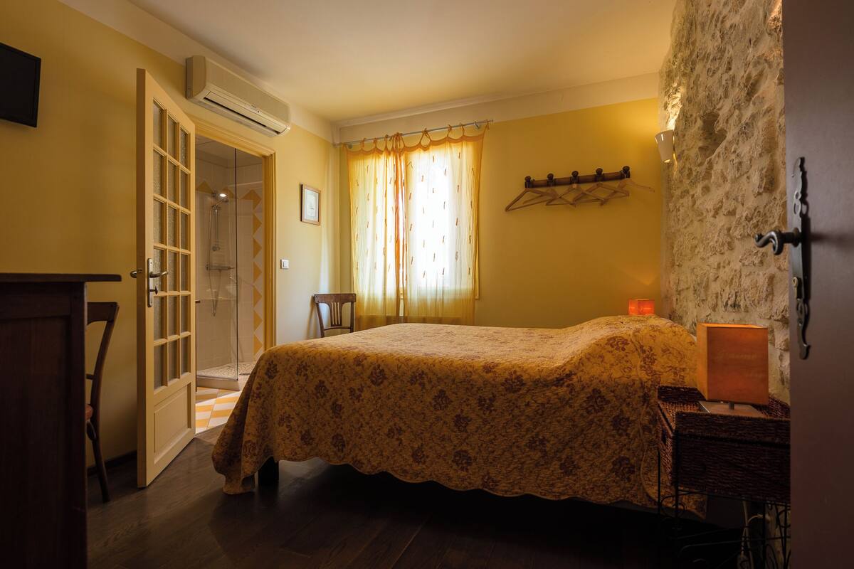 A bedroom with a bed, a chair, a desk, a lamp, and a wall-mounted shelf.