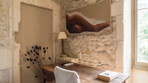 Una habitación rústica con pared de piedra, un escritorio de madera, una silla blanca, una lámpara y un cuadro de una figura reclinada.