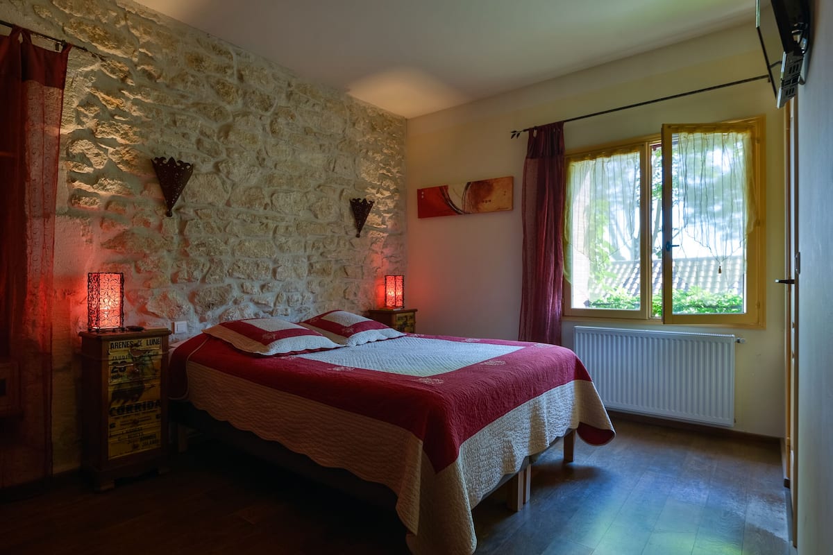 A bedroom with a stone wall, a bed with a red and white bedspread, two wall-mounted lamps, a window with curtains, and a radiator.