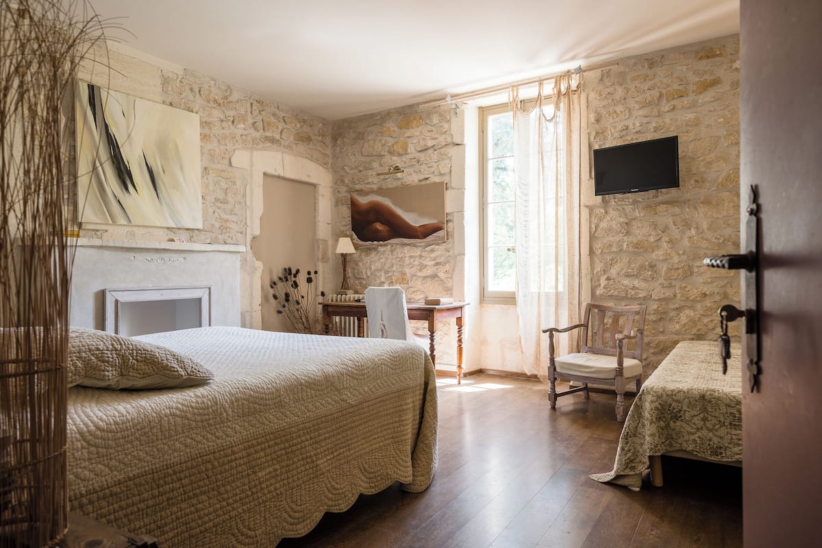 A bedroom with a stone wall, a bed with a quilt, a chair, a desk, and a TV mounted on the wall.