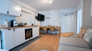 Une cuisine moderne avec des armoires blanches, un four encastré et une machine à café. On y trouve un coin repas avec une table et des chaises, ainsi qu’un salon avec un canapé et un téléviseur fixé au mur.