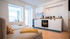 Un appartement moderne doté d’une kitchenette, d’un canapé agrémenté d’un plaid et d’une grande fenêtre laissant entrer la lumière naturelle.