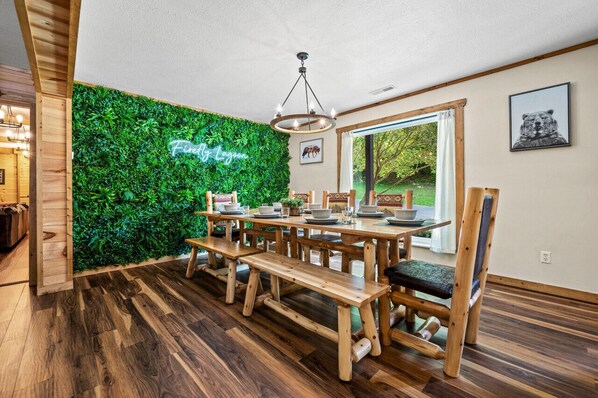 A dining area with a wooden table and benches, a green wall with a neon sign, and a window with a view of greenery.