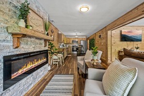 A cozy living room with a fireplace, wooden flooring, and a kitchen in the background.