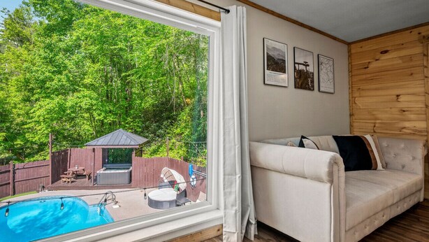 Ein Zimmer mit einem großen Fenster, das auf einen Garten mit Whirlpool, Terrasse und Pool blickt. Es gibt ein weißes Sofa und eine Holzverkleidung an der Wand.