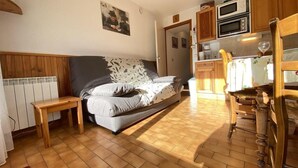 Une pièce confortable comprenant un lit, une petite table en bois, un coin cuisine avec des placards en bois et un four à micro-ondes, ainsi qu’un radiateur.