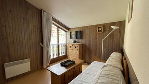 A small, cozy room with wooden walls, a bed, a desk with a chair, a TV, and a window with curtains.