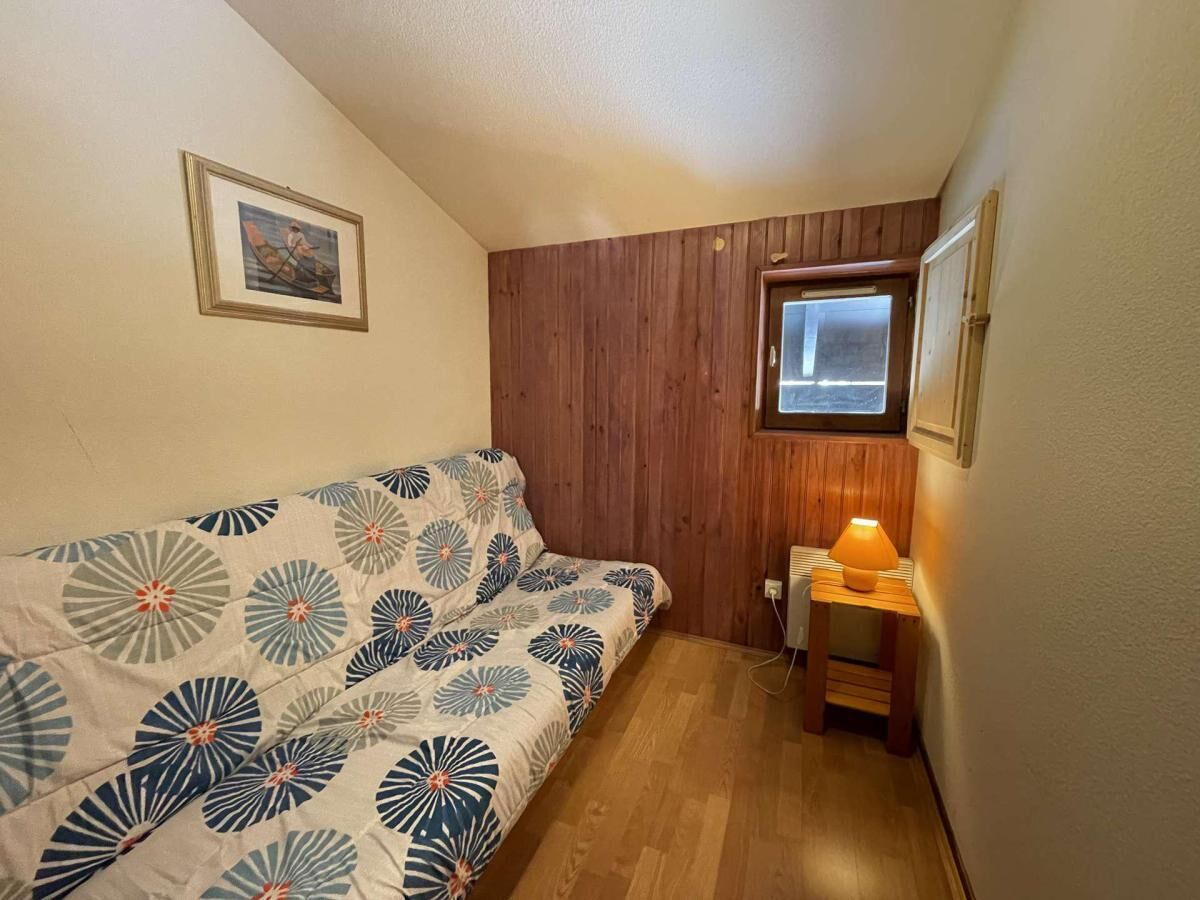 A small, cozy room with a wooden wall, a single bed with a floral bedspread, a small table with a lamp, and a window with a view.
