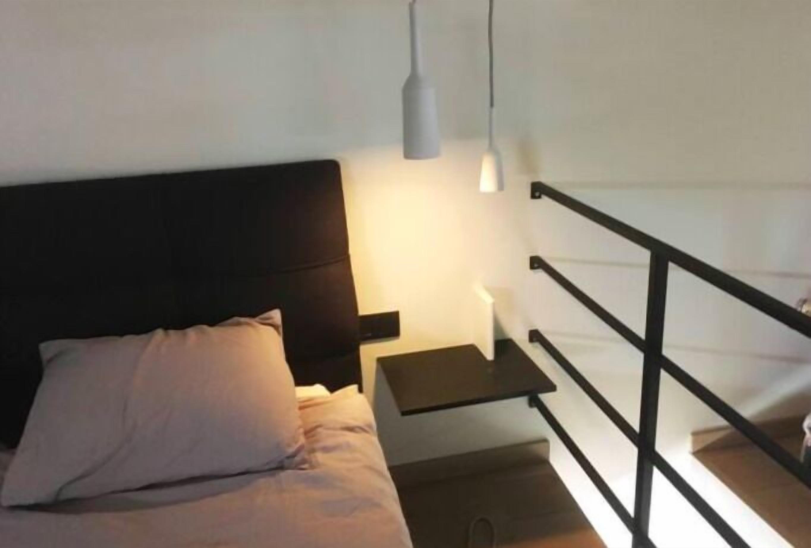 Een slaapkamer met een bed, nachtkastje, een wandplank en een zwarte metalen leuning.