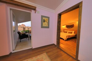 Quarto de hotel com cama, cadeira, mesa pequena e vista para uma paisagem montanhosa através de uma porta aberta.