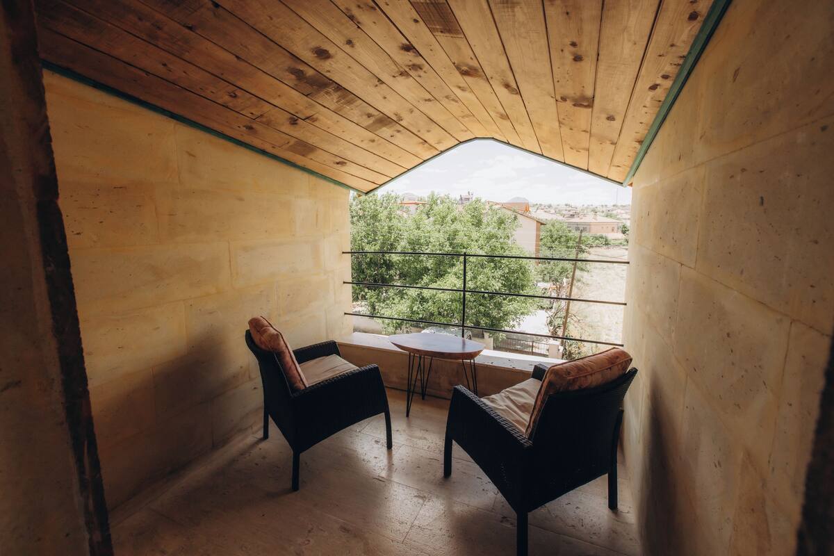 A small balcony with two wicker chairs and a table, wooden walls, and a view of greenery and buildings.