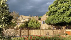 Un bâtiment rustique en pierre, doté d’un toit à pente traditionnelle, entouré d’une végétation luxuriante et d’une clôture en métal.
