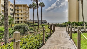 Une promenade en bois mène à un hôtel situé en bord de mer, bordé de palmiers, sous un ciel dégagé.