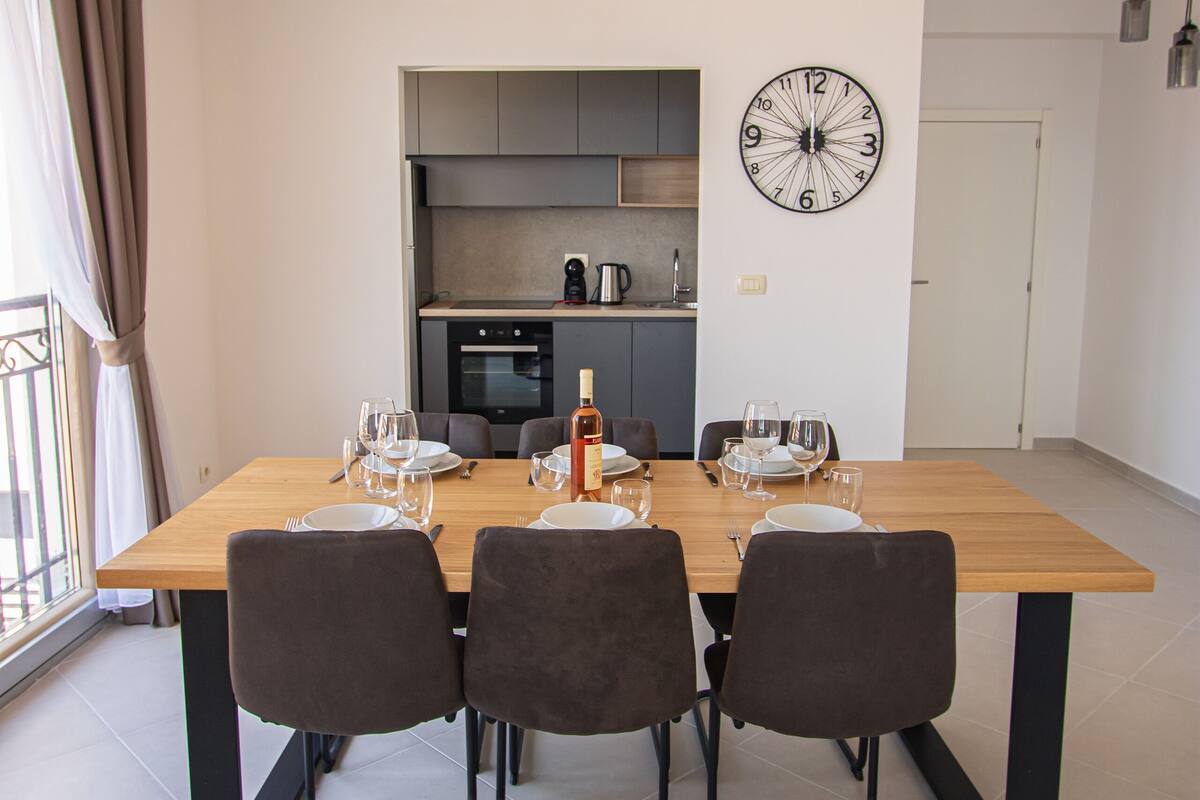 Un coin repas avec une table en bois dressée pour quatre personnes, une cuisine en arrière-plan et une grande horloge murale.