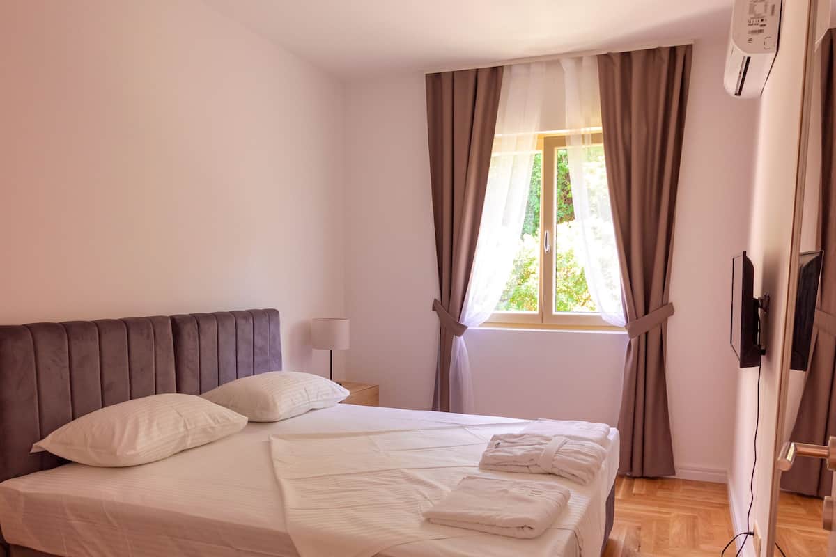 Un lit bien fait, avec du linge de lit blanc et une serviette pliée, une lampe de chevet et une fenêtre avec des rideaux.