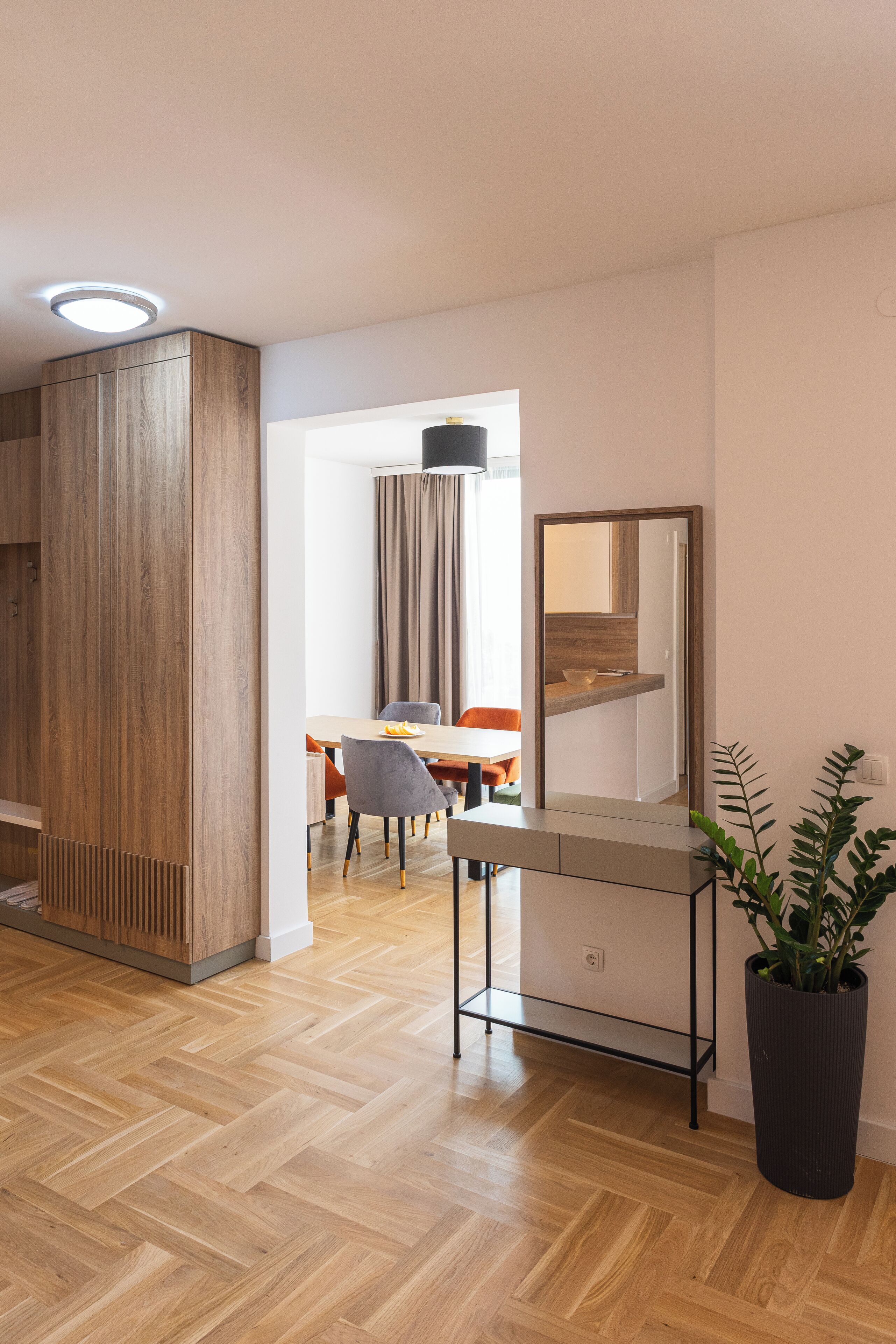 Un interior moderno con piso de madera, una zona de comedor con mesa y sillas, un espejo grande y una planta en maceta.