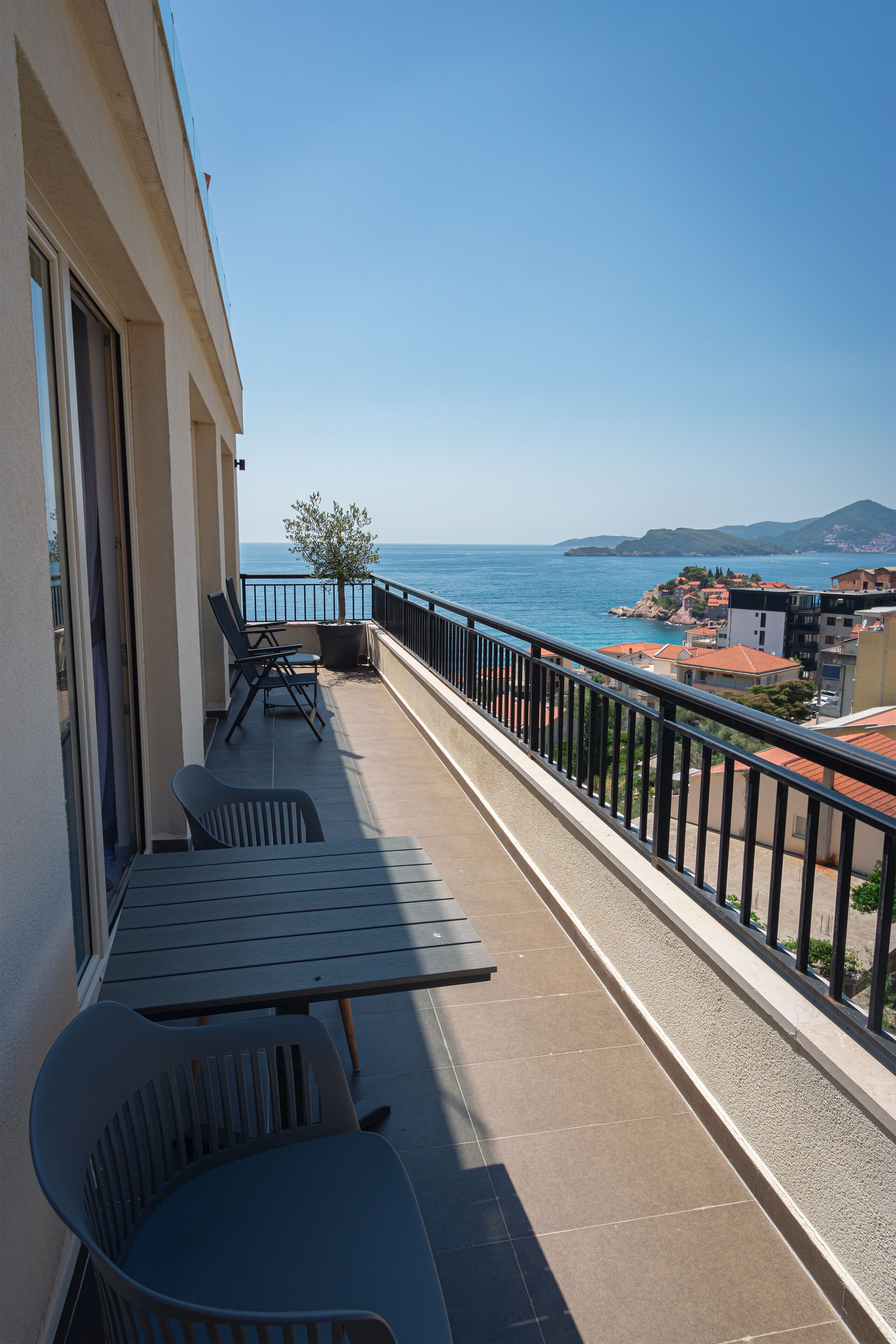 A balcony with a table and chairs, a potted plant, and a view of the sea and buildings.