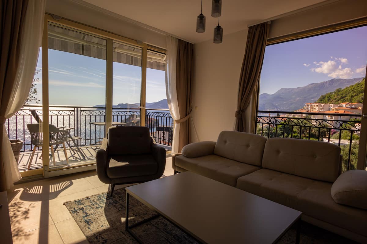 Un salon comprenant un canapé, un fauteuil et une table. Il est doté de grandes fenêtres offrant une vue sur un balcon et des montagnes.