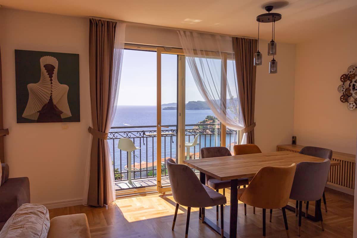 Un espace repas avec une grande table et des chaises, une vue sur la mer depuis un balcon, et un tableau accroché au mur.