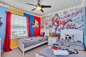 Quarto infantil com duas camas, um ventilador de teto e uma janela com cortinas vermelhas.