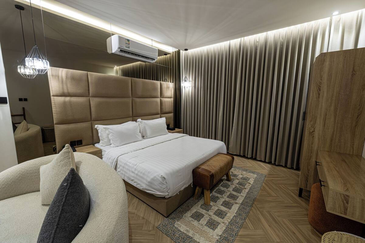 A modern hotel room with a large bed, a headboard, a bench, a small table, and a hanging light fixture.