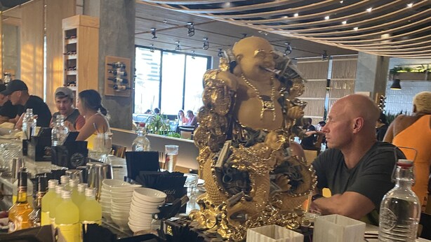 Ein Restaurantinnenraum mit Tischen, Stühlen und verschiedenen Gegenständen auf den Tischen, darunter Flaschen, Gläser und eine große, dekorative goldene Statue.