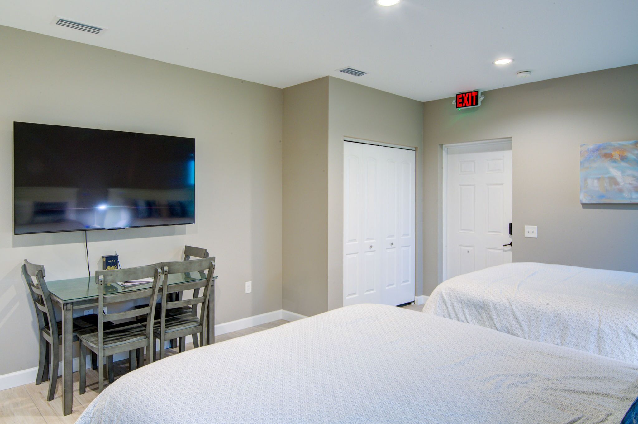 A hotel room with a TV, a dining table with chairs, two beds, and an exit sign.