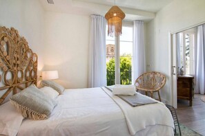 A bedroom with a large bed, a wicker chair, a hanging light fixture, and a view of greenery outside. - Alhambra Luxviews Apartment (Granada)