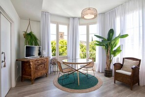 A room with a wooden desk, a television, a round table with chairs, a large potted plant, and a wicker chair.
