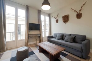 A living room with a sofa, a coffee table, and a television. There are decorative wall pieces and a view through the window.