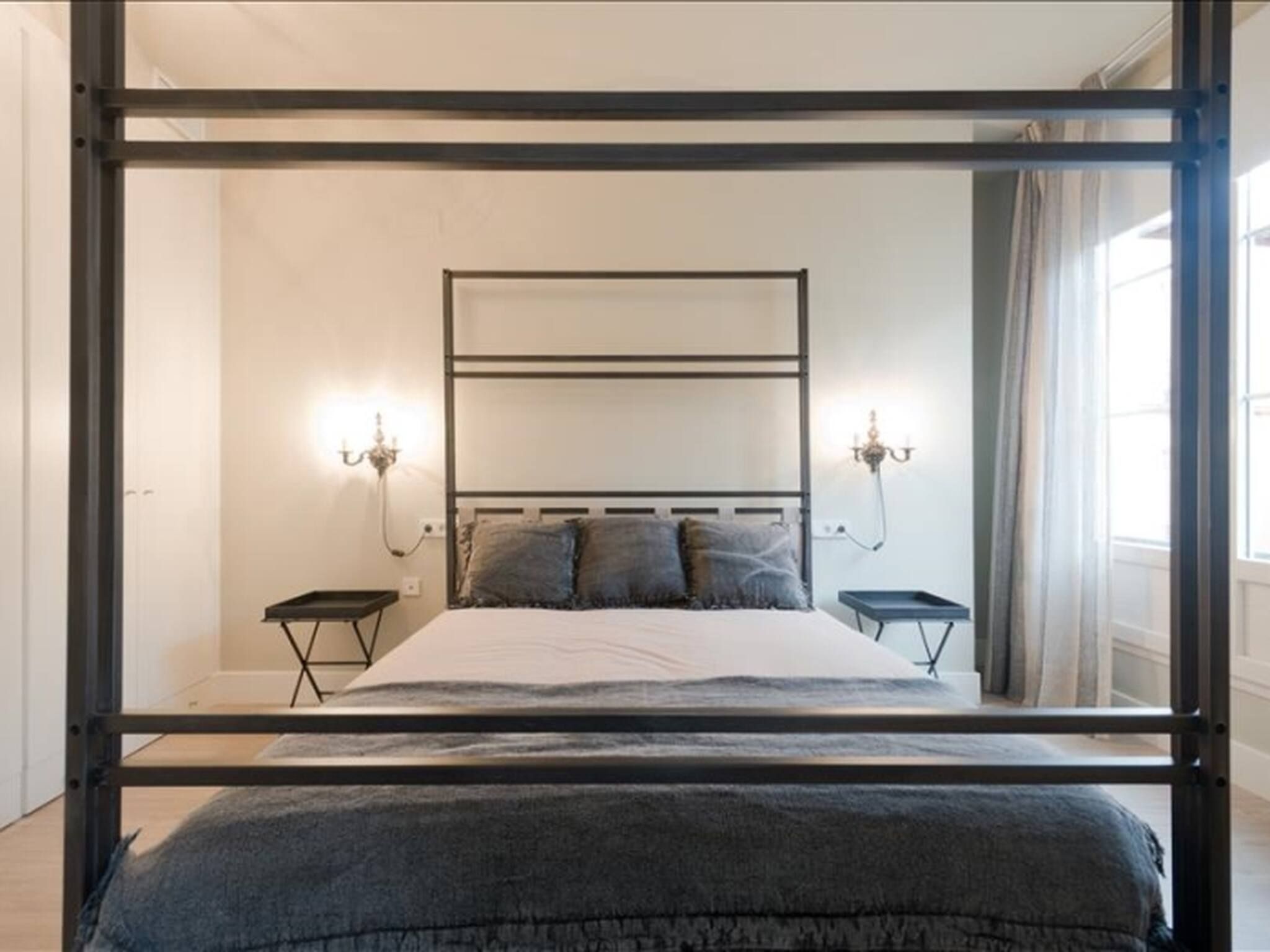Een slaapkamer met een bed met metalen frame, twee nachtkastjes, een wandlamp en een raam met gordijnen.