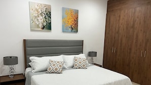 A bedroom with a grey upholstered headboard, white textured bedspread, and decorative pillows. There are two bedside tables with lamps, a wooden wardrobe, and two framed floral paintings on the wall. - New & beautiful apartment (GF) with private pool (Mérida)