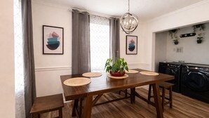Un coin repas avec une table en bois, des sets de table en fibres tressées, un luminaire suspendu et une vue sur une buanderie équipée de machines à laver.