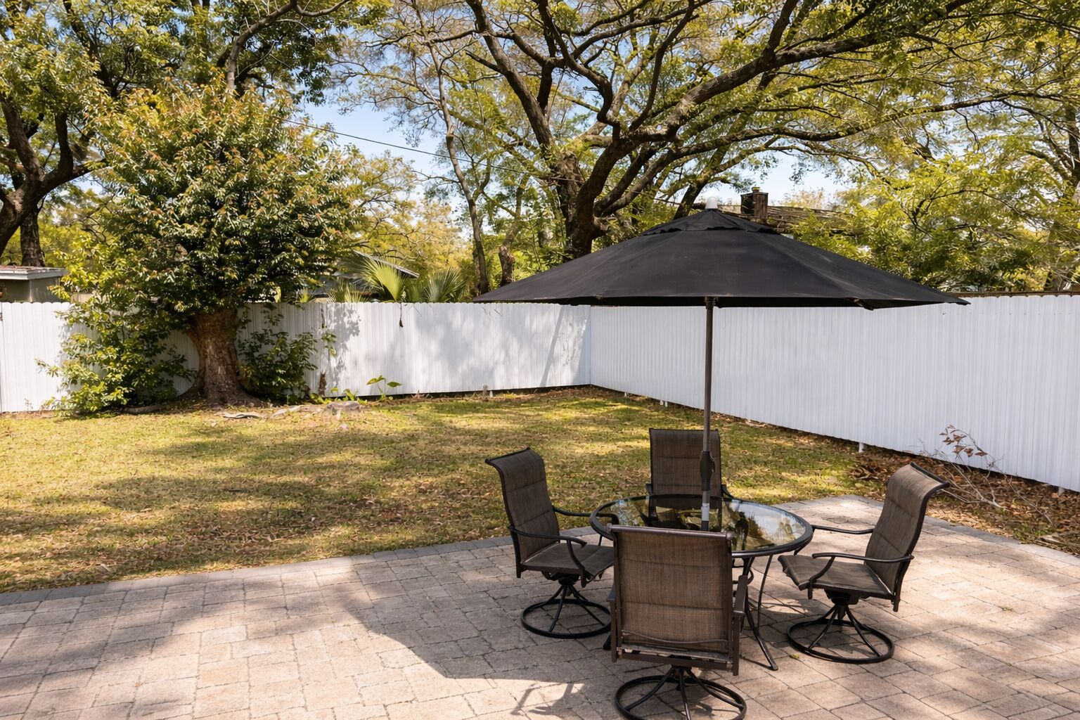 An outdoor patio with a table and chairs, a large umbrella, and a white fence in the background.