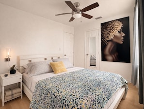 A bedroom with a bed, a ceiling fan, a nightstand, a wall-mounted lamp, and a large framed picture.