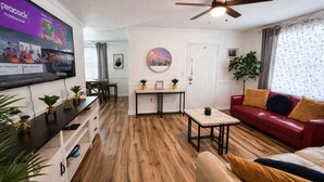 Un salon avec un canapé rouge, une table basse, un téléviseur fixé au mur, un ventilateur de plafond et un coin repas avec une table et des chaises.
