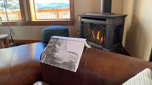 A cozy living room with a brown leather couch, a fireplace, and a view of a wooden deck through a large window.