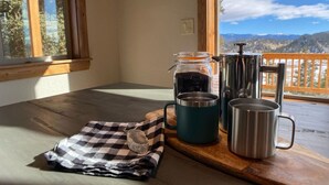 A wooden table with a checkered cloth, a glass jar, and stainless steel mugs on a balcony with a mountain view.