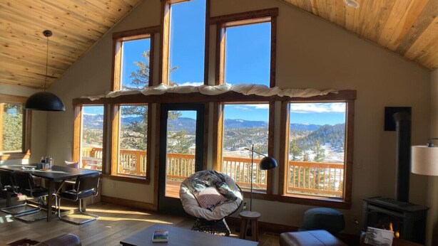 A cozy living room with a high ceiling, large windows offering a view of a mountainous landscape, a dining area with a table and chairs, and a fireplace.