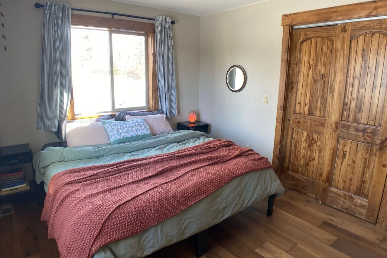 Un dormitorio con una cama, una ventana con cortinas, una puerta de madera y una mesita de noche con lámpara.