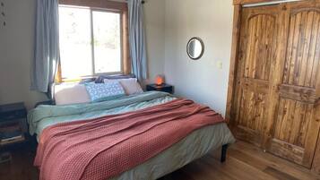 Un dormitorio con una cama, una ventana con cortinas, una puerta de madera y una mesita de noche con lámpara.