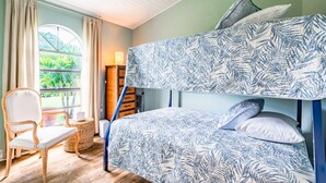 Una cama litera con una colcha estampada con motivos de hojas, una silla de mimbre y vistas al verdor a través de una ventana.