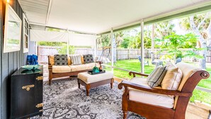 Un área de estar al aire libre con techo, que incluye un sofá, un reposapiés y una silla de madera, un aparador oscuro y una alfombra estampada.