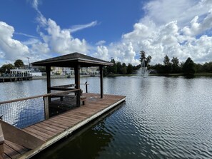 A wooden dock extending into a calm lake, with a gazebo and a fountain in the background.