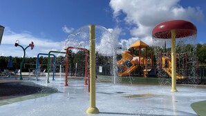 Un espace de jeux aquatiques doté de plusieurs jets d’eau et, au fond, une structure de jeux pour enfants.