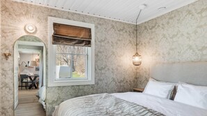 A bedroom with a large bed, a window with a brown blind, a wall-mounted light fixture, and a door leading to a dining area.