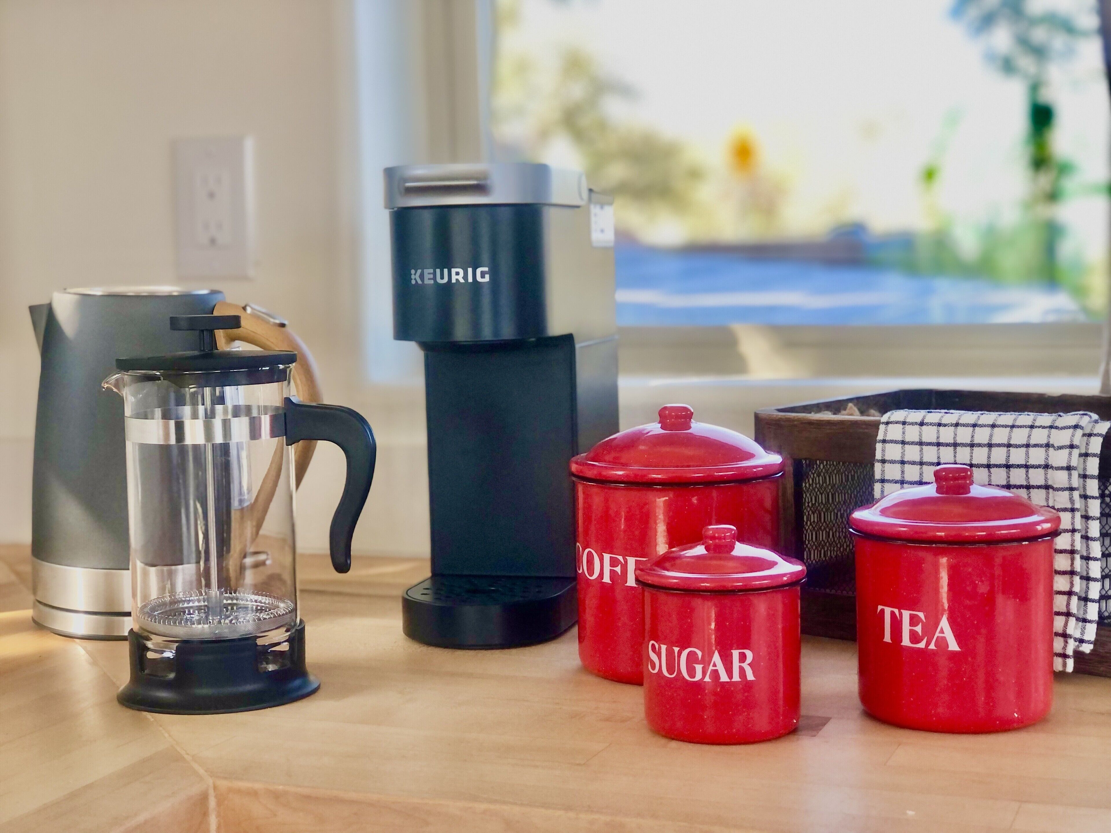 Una encimera de cocina con una cafetera, un hervidor y dos recipientes rojos etiquetados 'SUGAR' y 'TEA'.