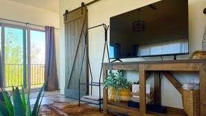 Una sala de estar con un televisor grande, un mueble de televisión de madera, una puerta corrediza de estilo rústico, un tapete de cuero de vaca y un balcón con vistas a árboles.