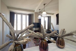 A modern living room with a dining area, featuring a large window, a flat-screen TV, and decorative wooden branches in vases.