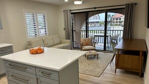A modern kitchen with a central island, a dining area with a wooden table, and a view of the street through large windows. - Sunshine Swimmers Casita, 206 (Englewood)